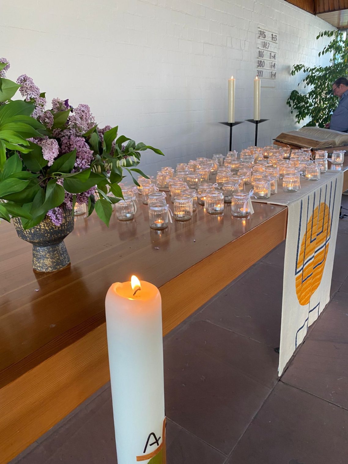 Gedenkgottesdienst am 29. April 2026 Tisch mit Kerzen, Blumenarrangement und Banner in einem hellen Raum.