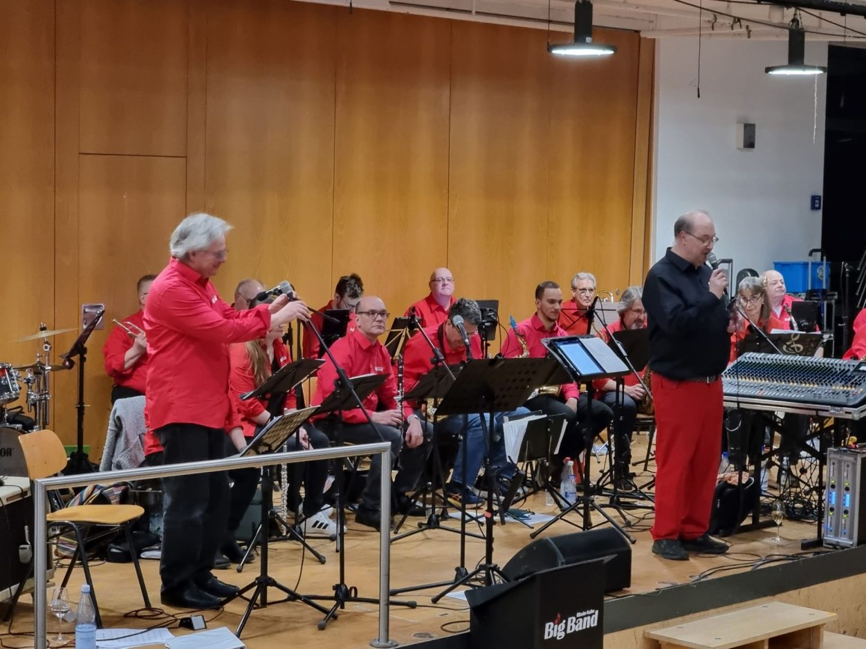 Benefizkonzert der Rhein-Nahe Big Band Eine Kapelle tritt in roten Outfits auf, während der Dirigent das Orchester anleitet.