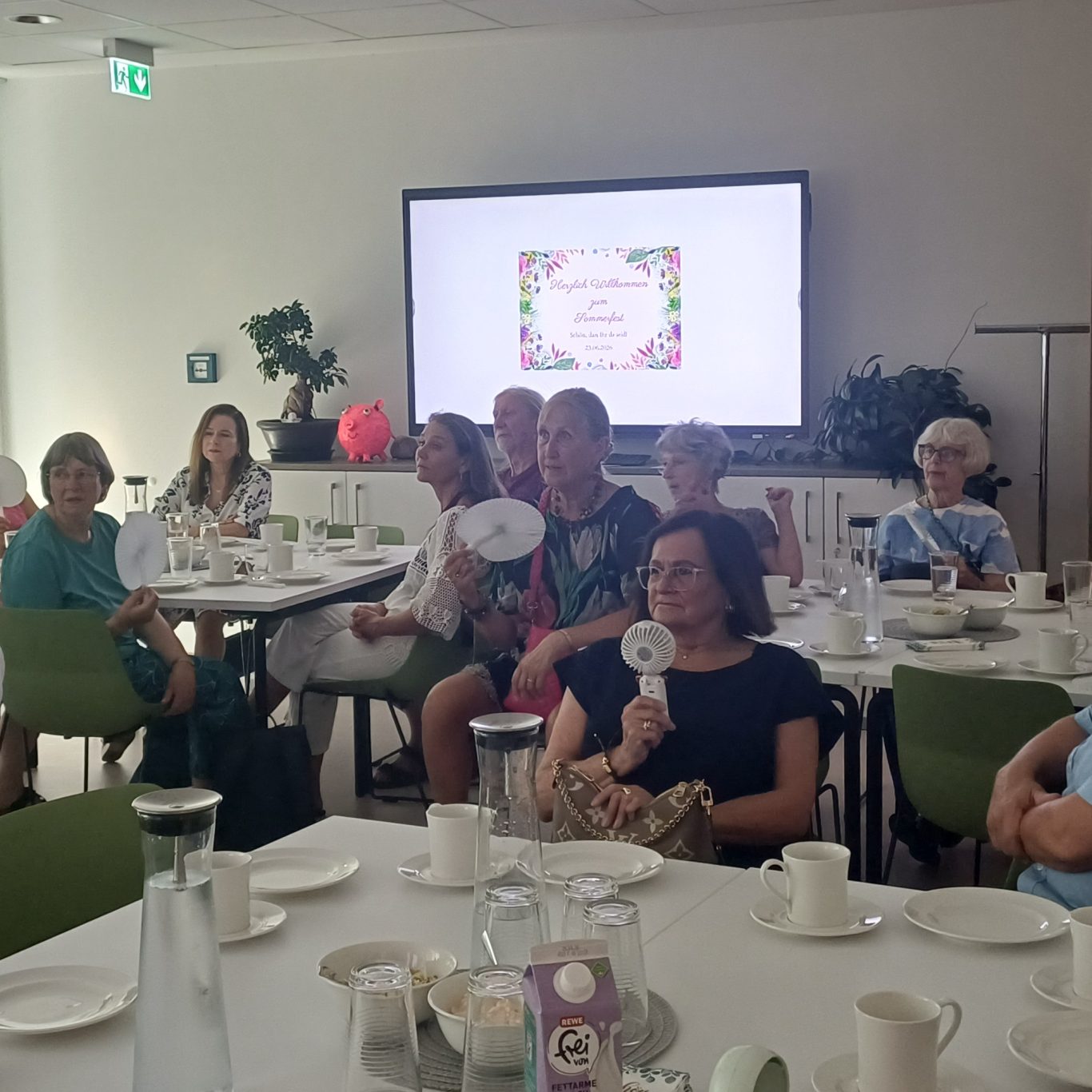 23. Juni 2026 Sommerfest für unsere ehrenamtlichen Helferinnen und Helfer Eine Gruppe von Menschen sitzt an einem Tisch und hält Fächer während einer Veranstaltung.
