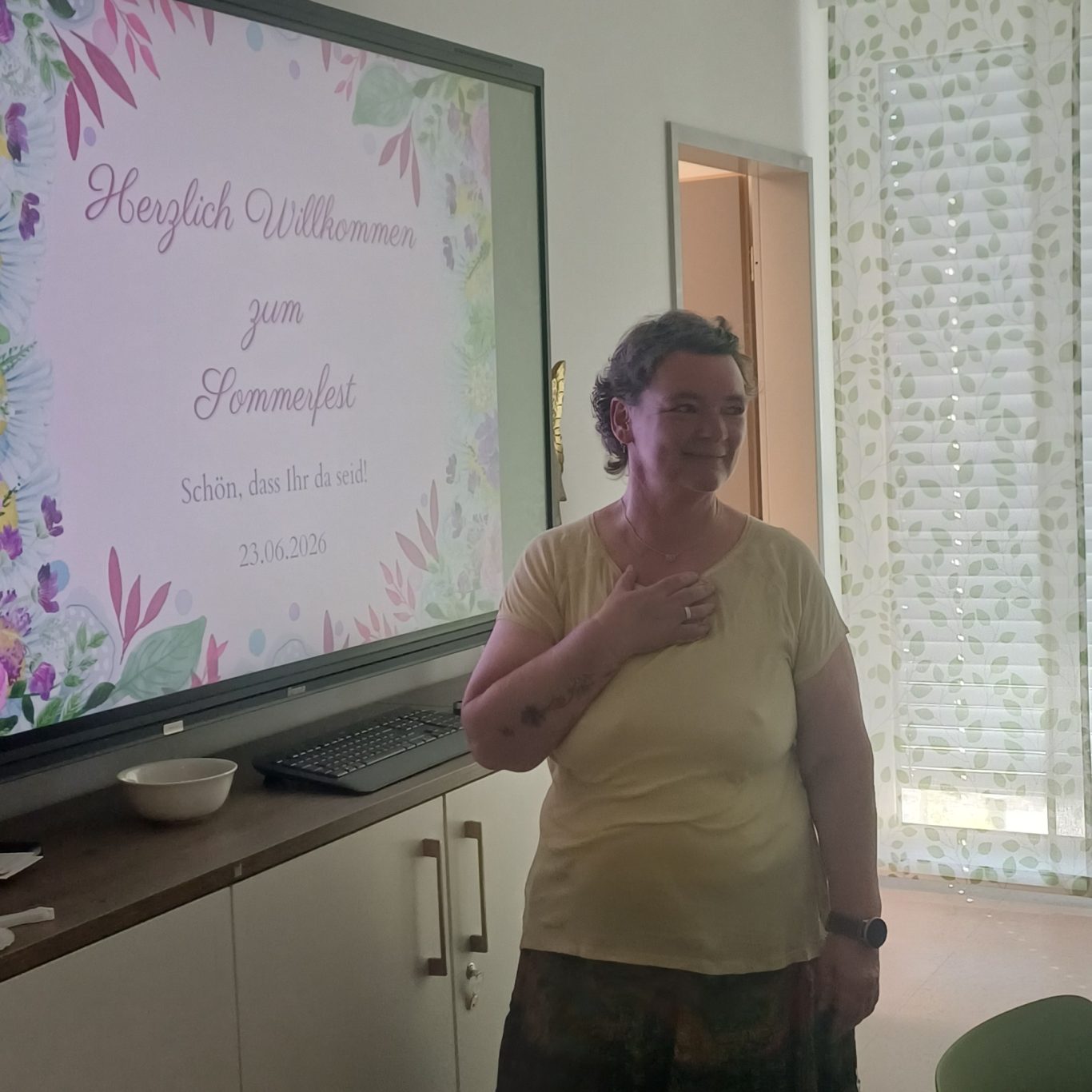 23. Juni 2026 Sommerfest für unsere ehrenamtlichen Helferinnen und Helfer Bianca Meyer steht an einer Wand mit der Einladung und lächelt.