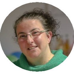 Eine lächelnde Frau mit lockigem Haar und Brille trägt einen grünen Pullover.