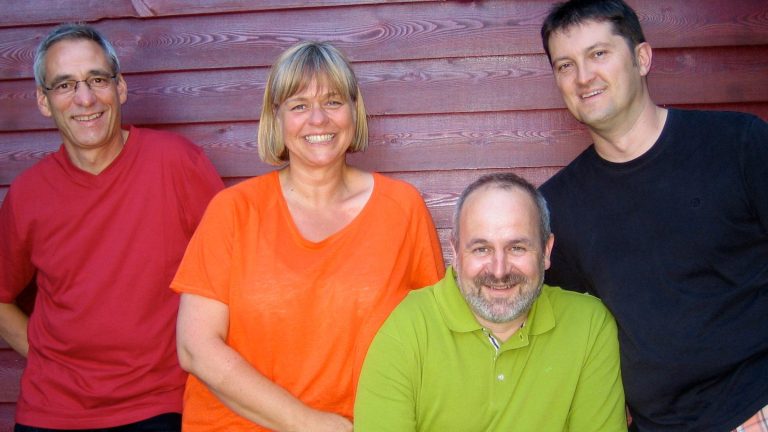 Vier Personen lächeln vor einer roten Wand, etwas unterschiedlich gekleidet.