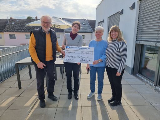 Vier Personen halten einen großen Scheck auf einer Terrasse bei Tageslicht.